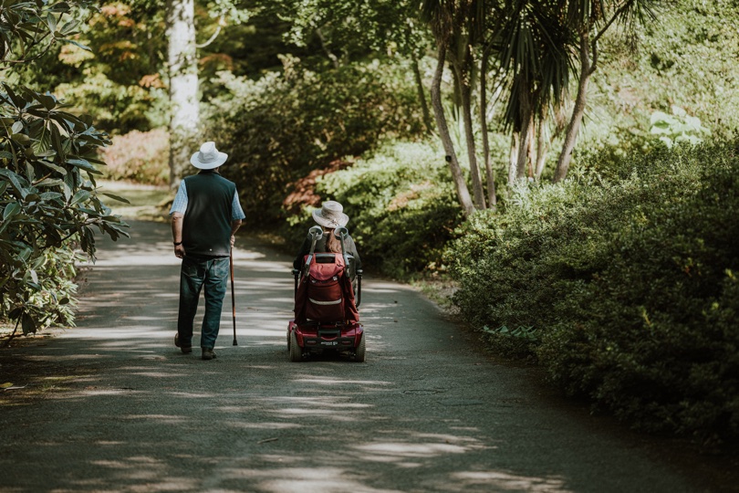 A man with a cane and a woman on a wheelchair walking down a dirt road