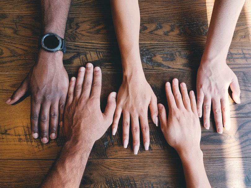 Five human hands of different skin colours on brown surface.