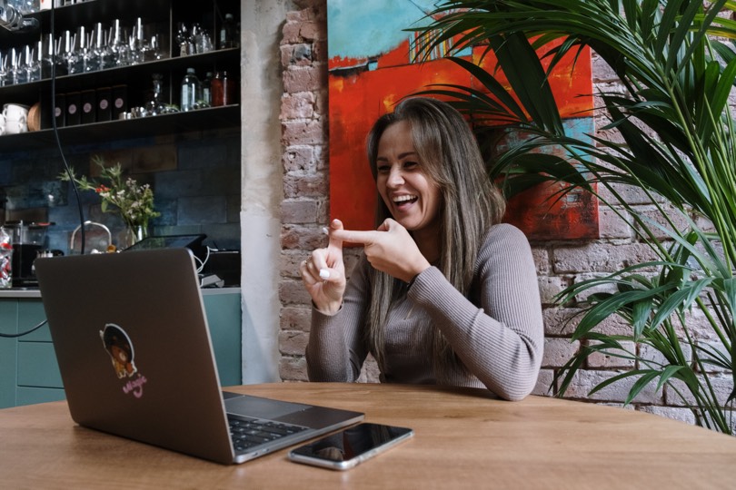 Woman sitting by a table doing hand sign while having video chat.