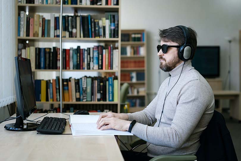 Homem em frente a um computador com uns auscultadores enquanto lê braille